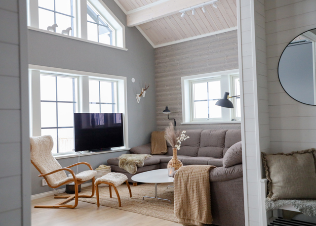 Ljust vardagsrum med hörnsoffa, fåtölj, TV och stora fönster.