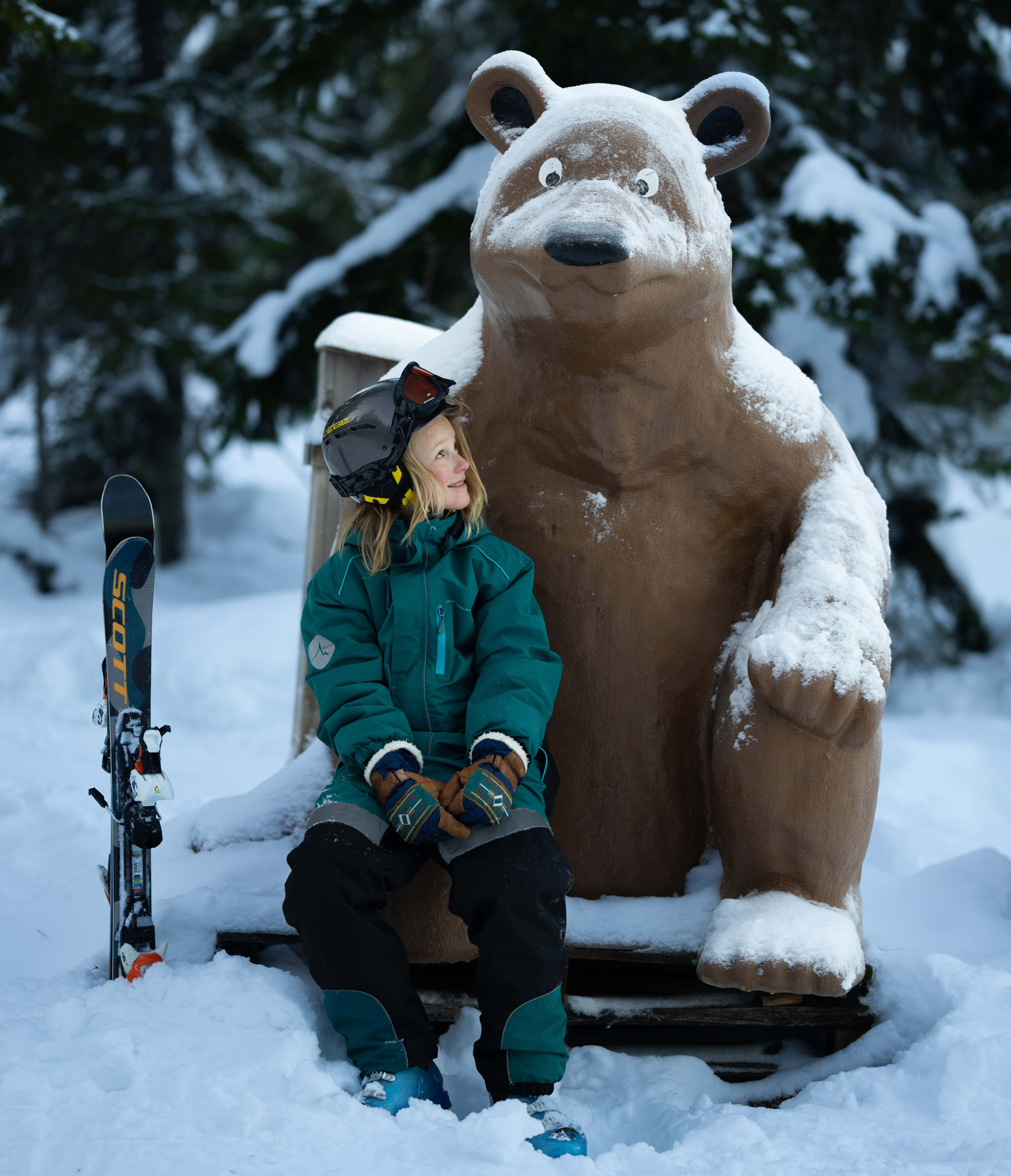 Barn i skidkläder sitter bredvid en snötäckt maskoten i vintrigt landskap.