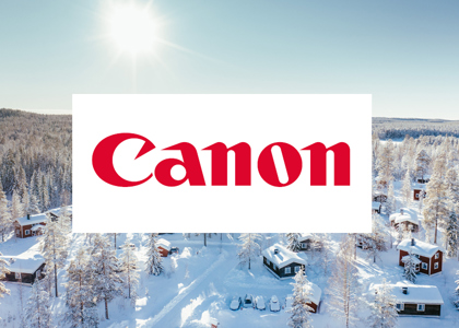 Logotyp för Canon centrerad över en solig vinterbild med snötäckta stugor och skog i bakgrunden.