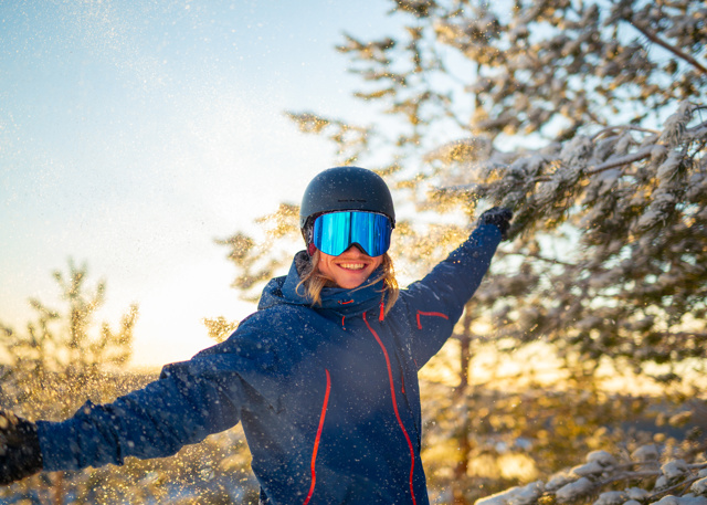 Smiling person in ski gear with arms outstretched, standing in a sparkling snowy landscape at sunset.