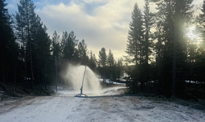 En snökanon som sprutar snö över ett frostigt fält i skogsmiljö, med tallar runtom och blek morgon- eller kvällssol bakom träden.