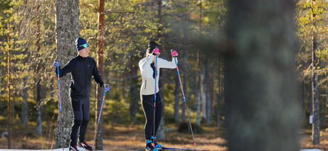 Två längdskidåkare på tur i ett solbelyst skogsparti med sparsam snö på marken, en i svart och en i vit jacka.