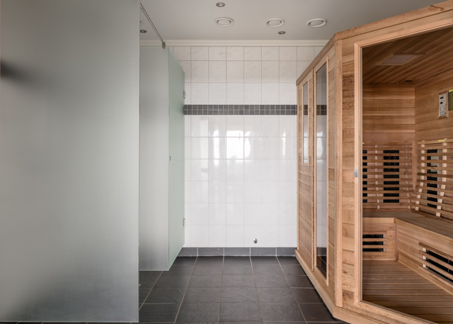 Badrum med vita kakelväggar, frostat glas som avskiljer duschutrymmen och en stor träbastu med glasdörr.