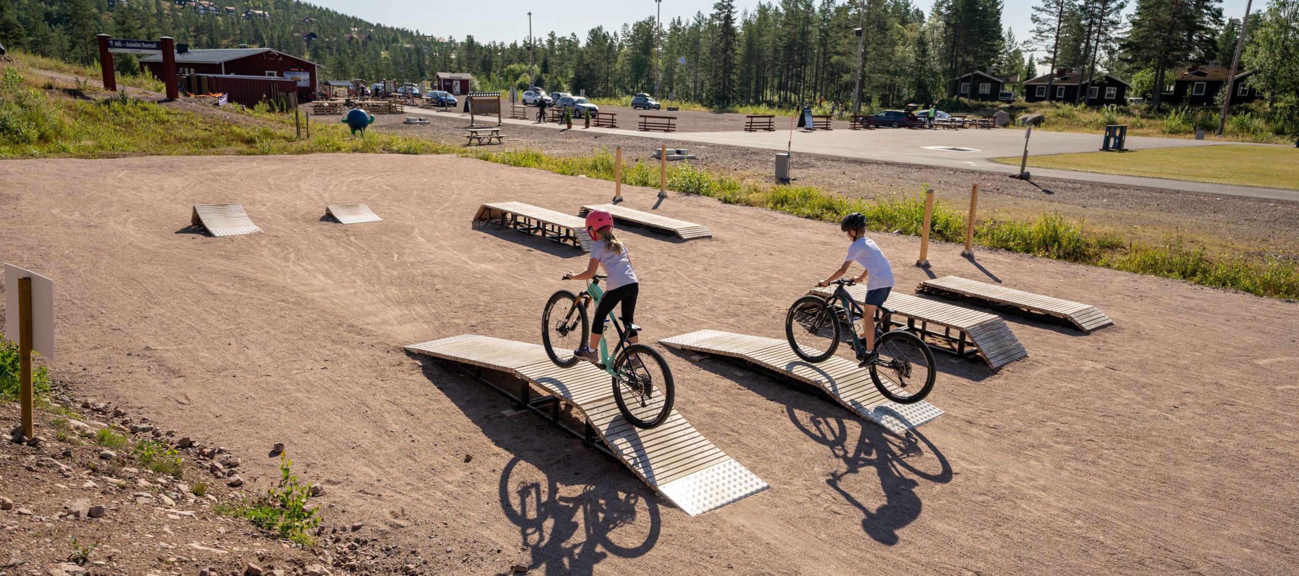 Två barn cyklar över små trähinder på en teknikbana för mountainbike, med grusunderlag och öppen yta intill skog och parkering i bakgrunden.