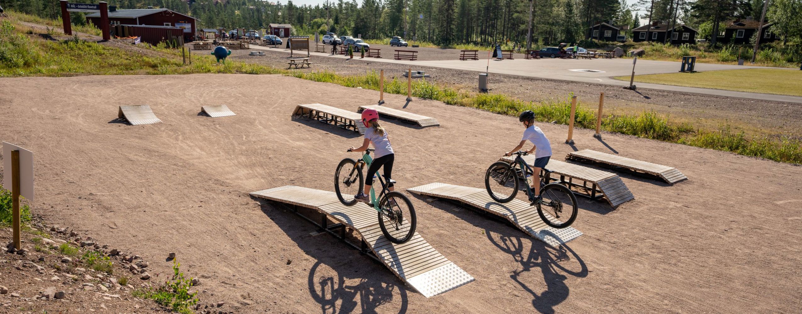 Two children biking over small wooden obstacles on a mountain bike skills course with gravel surface, near a forest and parking area in the background.
