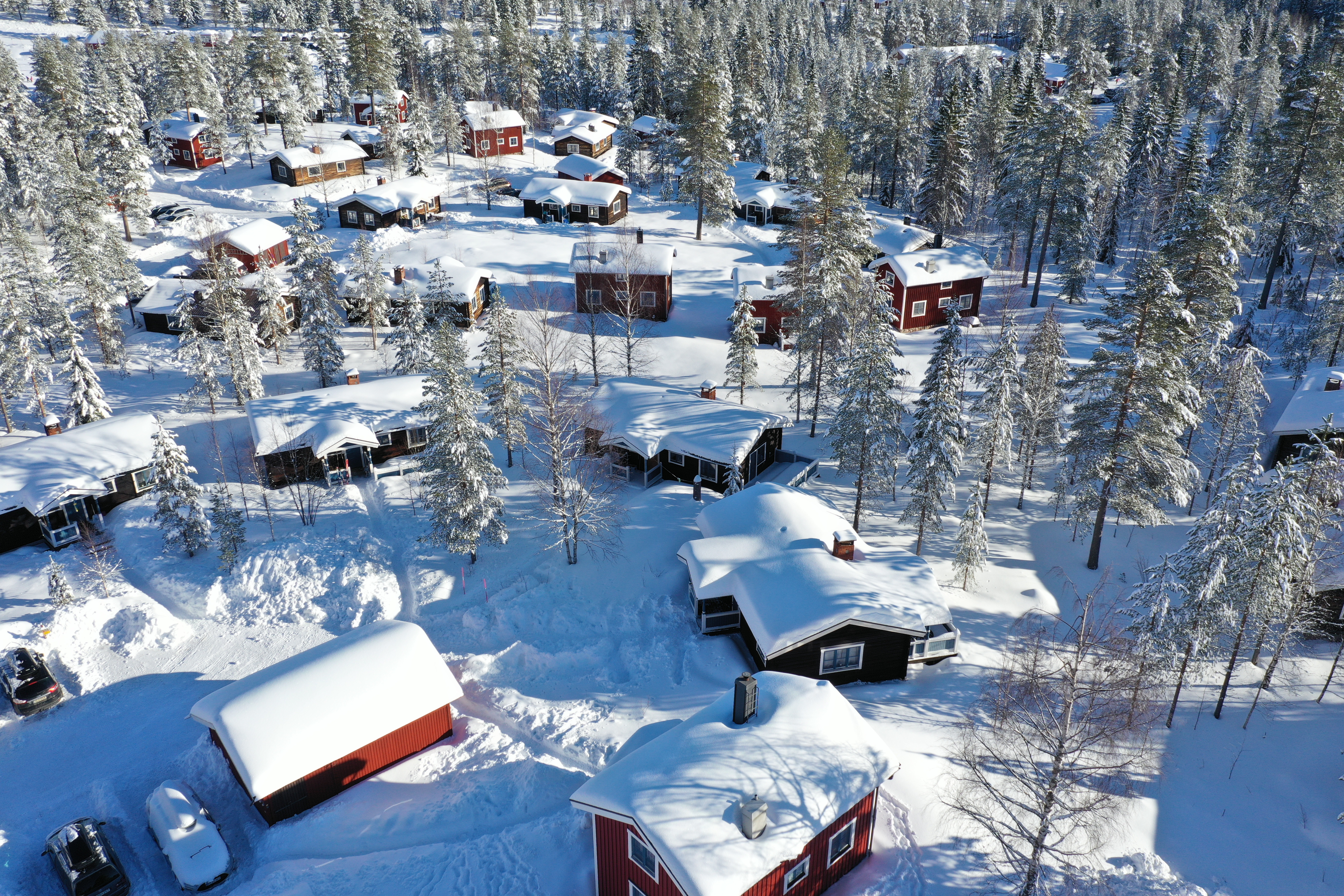 Aerial view of a snow-covered cabin area with red and dark cottages in a forest setting.