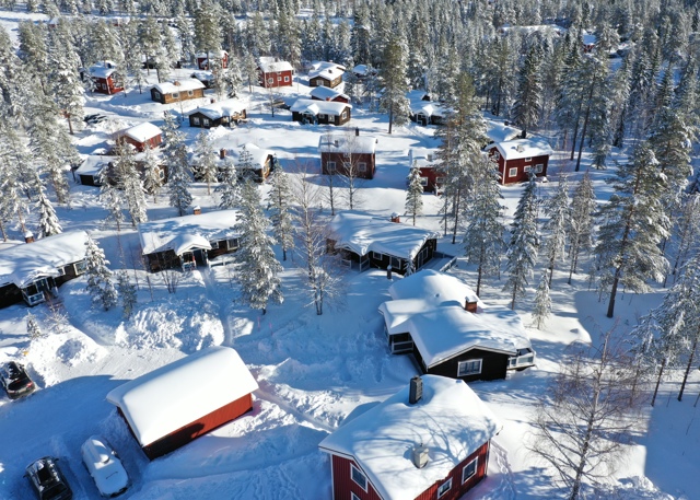 Flygfoto över snötäckt stugområde med röda och mörka stugor i skogsmiljö.