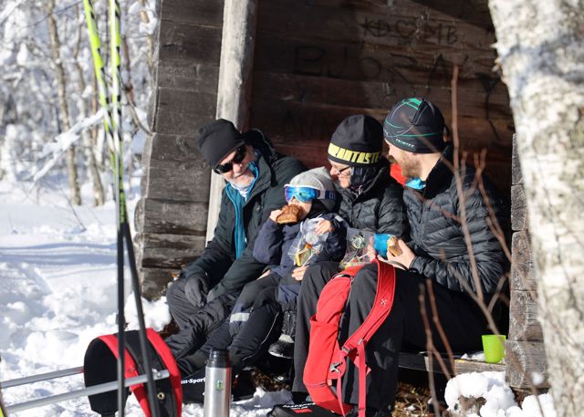 Familie i vintertøj holder pause med mad og drikke ved et læskur langs skiruten.
