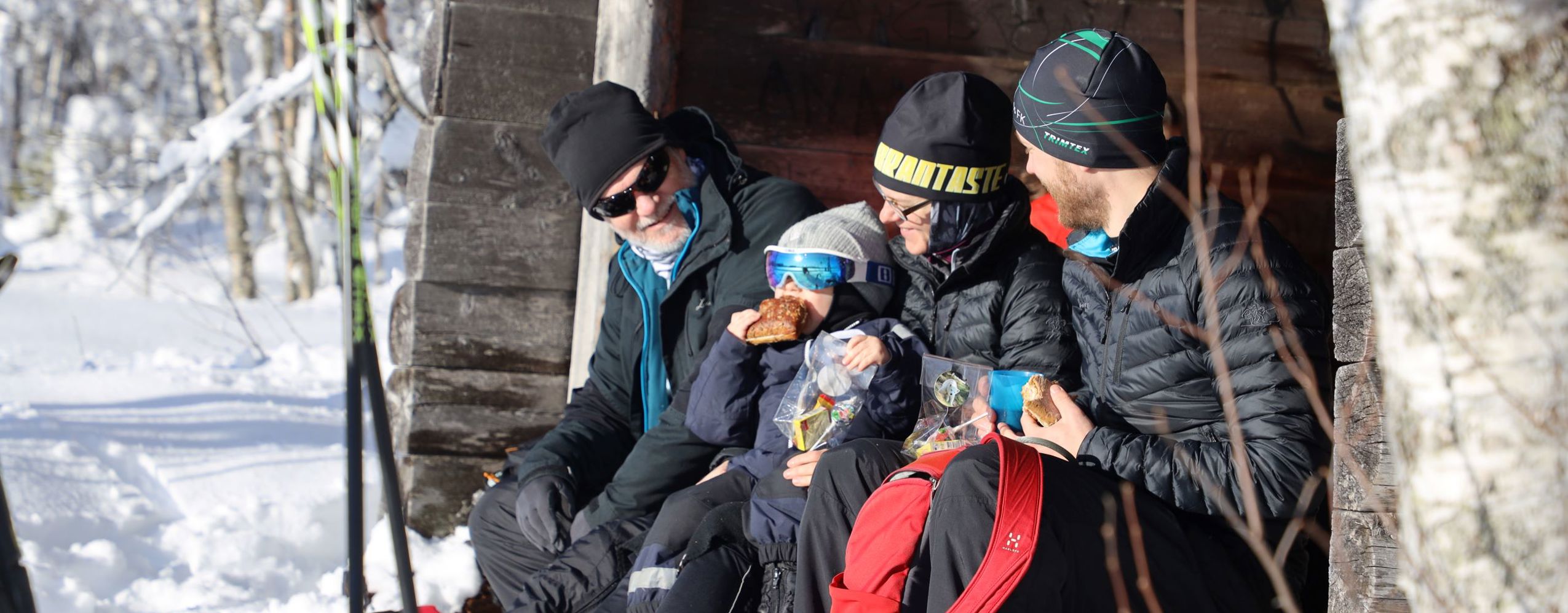 Familj i vinterkläder tar paus med fika vid ett vindskydd längs skidspåret.