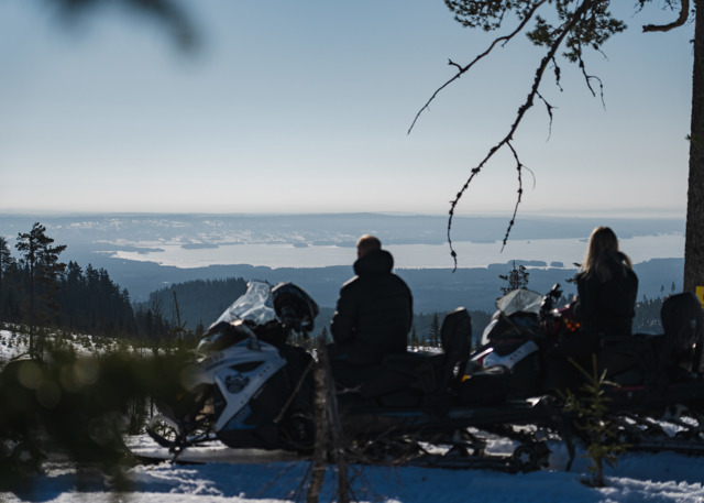 To personer på snescooter nyder udsigten over et sneklædt landskab og en sø i horisonten.