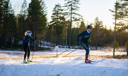 Två längdskidåkare tränar i soligt vinterlandskap omgivna av tallar och snö i Orsa Grönklitt.
