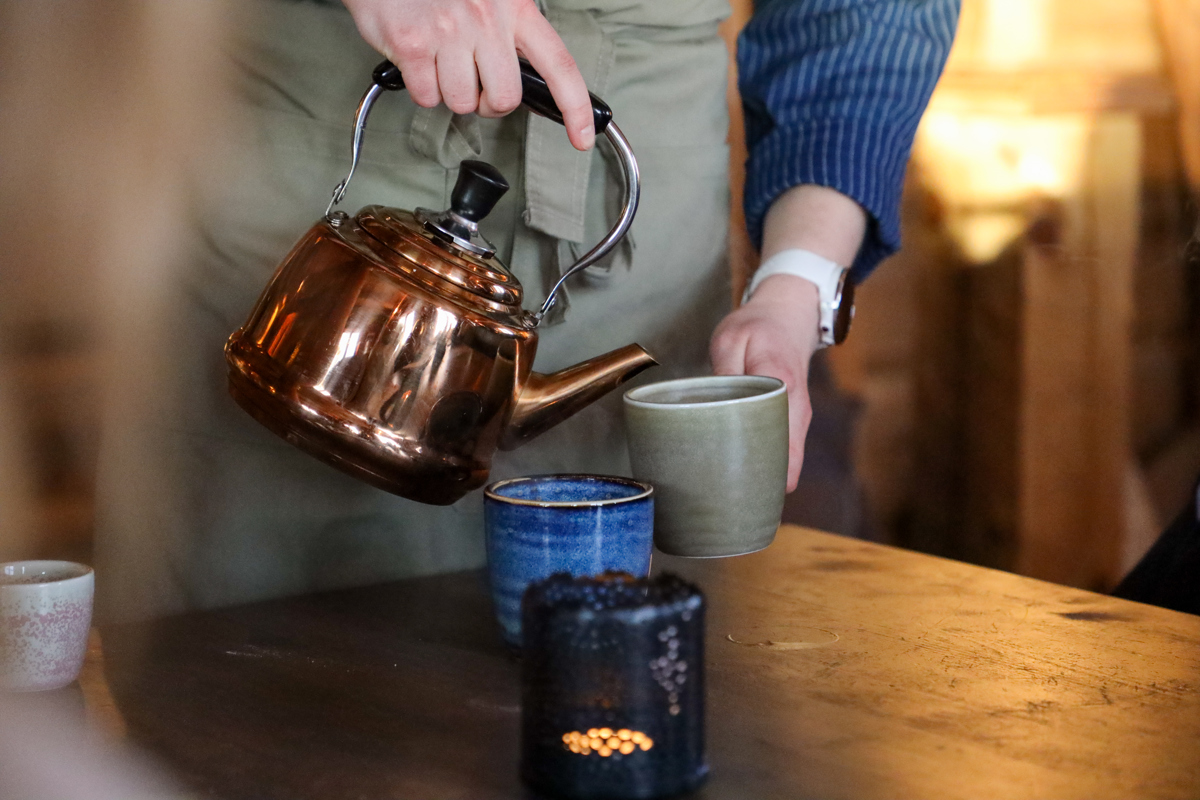 En person hælder en varm drik op fra en kobberfarvet tekande i en kop ved et rustikt bord i Orsa Grönklitt.