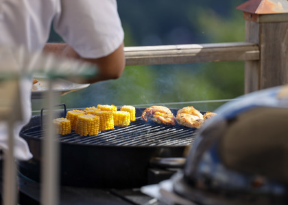 Nærbillede af en grill, hvor kylling og majskolber tilberedes udendørs.