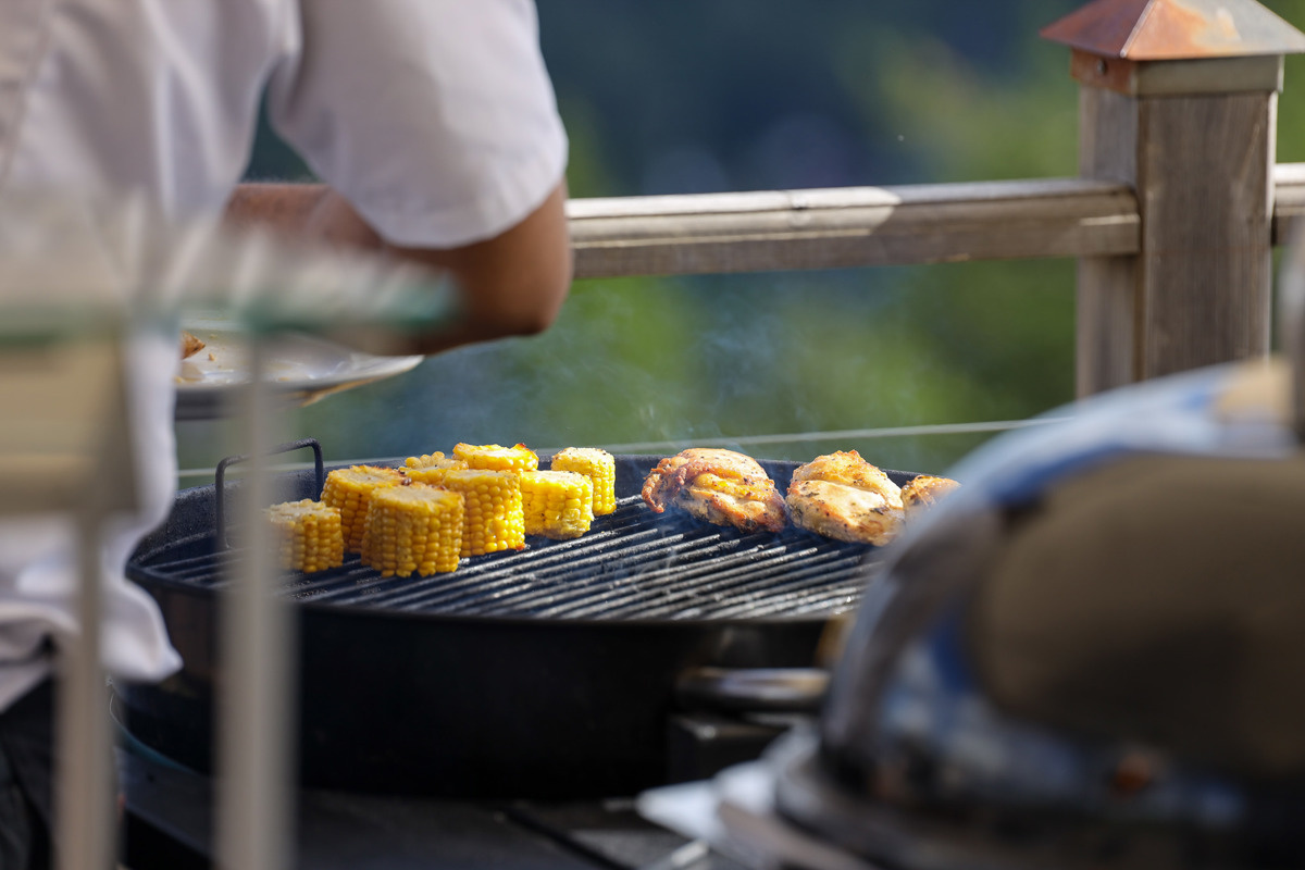 Nærbillede af en grill, hvor kylling og majskolber tilberedes udendørs.