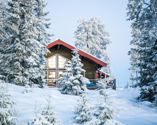Trästuga med stora fönster och rött tak i Orsa Grönklitt, omgiven av snötyngda granar.
