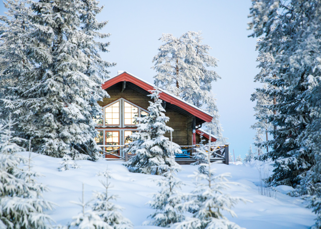 Wooden cabin with large windows and red roof at Orsa Grönklitt, surrounded by snow-covered pine trees.