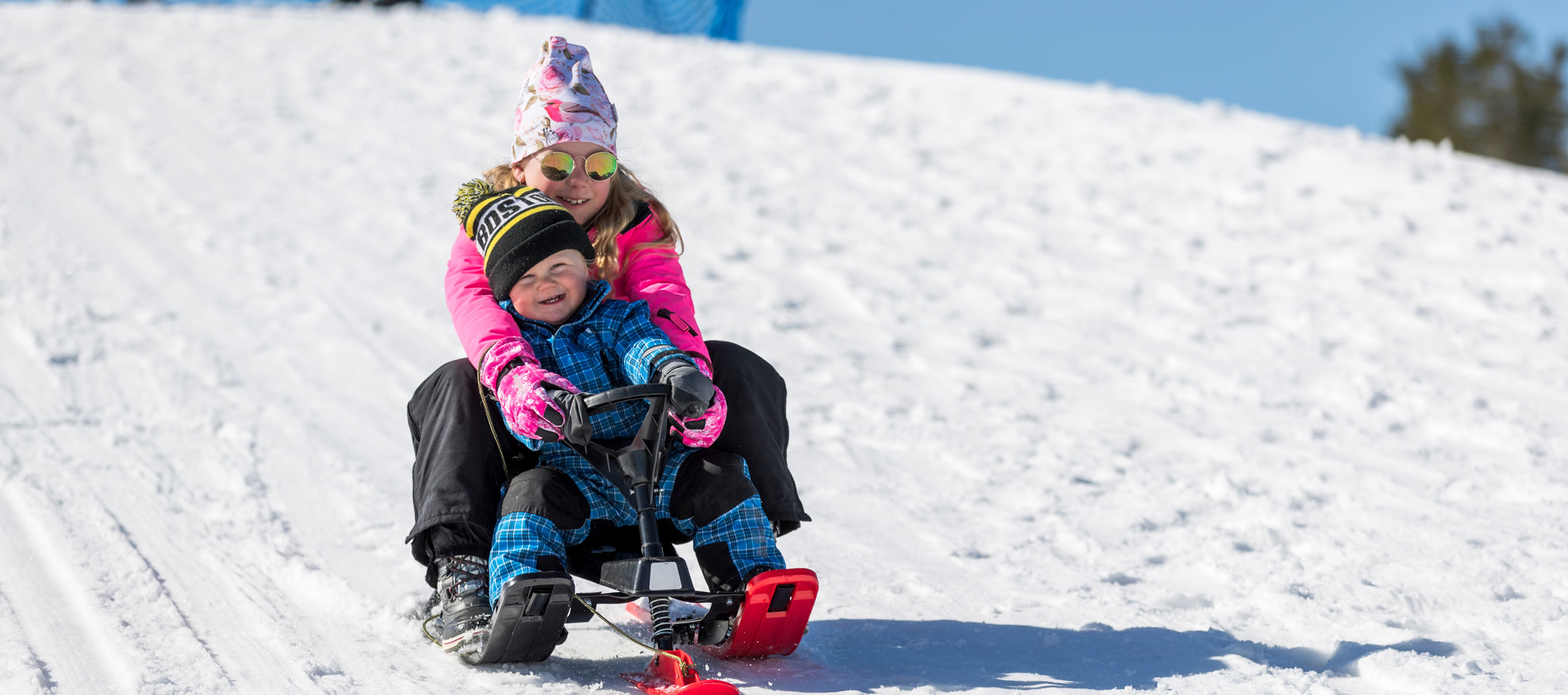 En flicka och ett barn åker snowracer nerför en snöig backe i solskenet. Båda ler stort.