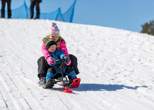 En flicka och ett barn åker snowracer nerför en snöig backe i solskenet. Båda ler stort.