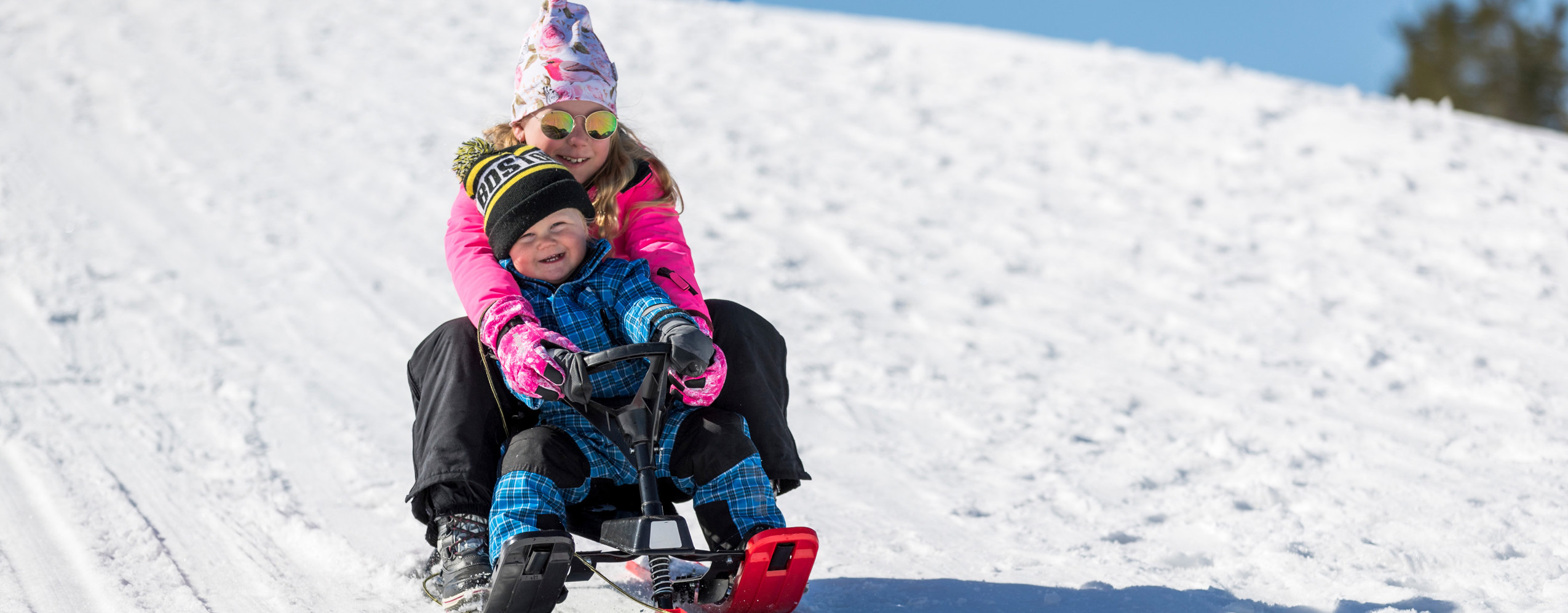 A girl and a young child are riding a snowracer down a snowy hill in the sunshine. Both are smiling brightly.