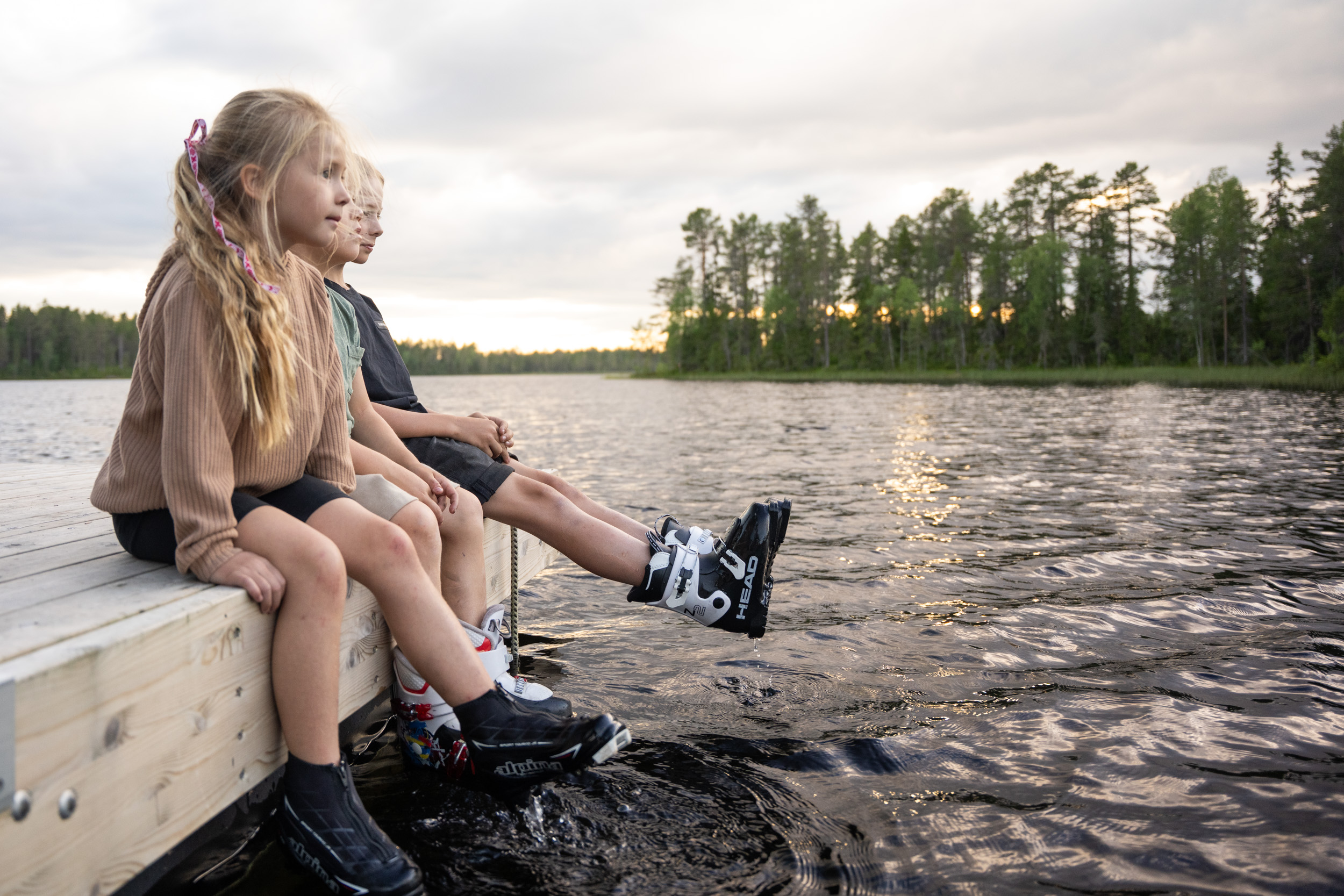 Tre barn sitter på en brygga med skidskor på fötterna och doppar dem i vattnet under en sommarkväll vid en skogssjö.