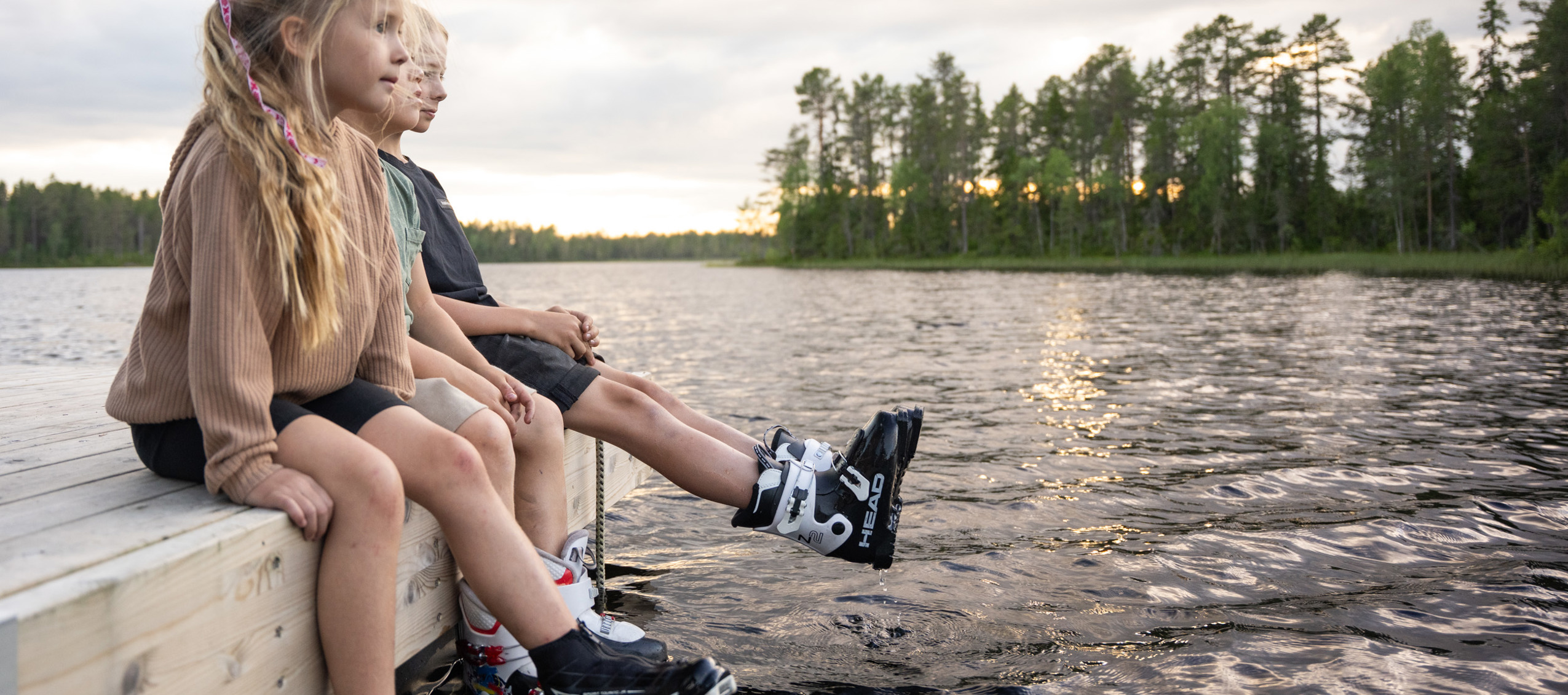 Tre barn sitter på en brygga med skidskor på fötterna och doppar dem i vattnet under en sommarkväll vid en skogssjö.