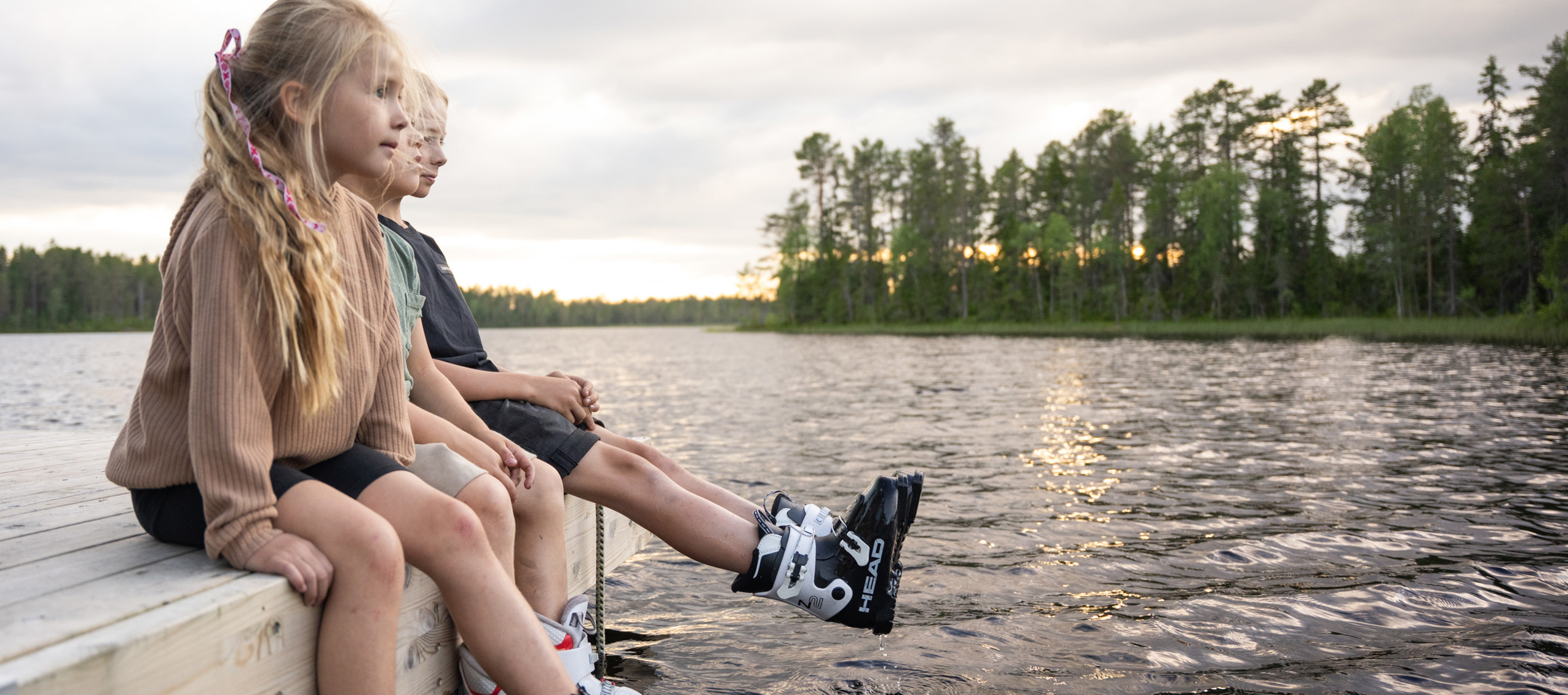 Tre barn sitter på en brygga med skidskor på fötterna och doppar dem i vattnet under en sommarkväll vid en skogssjö.