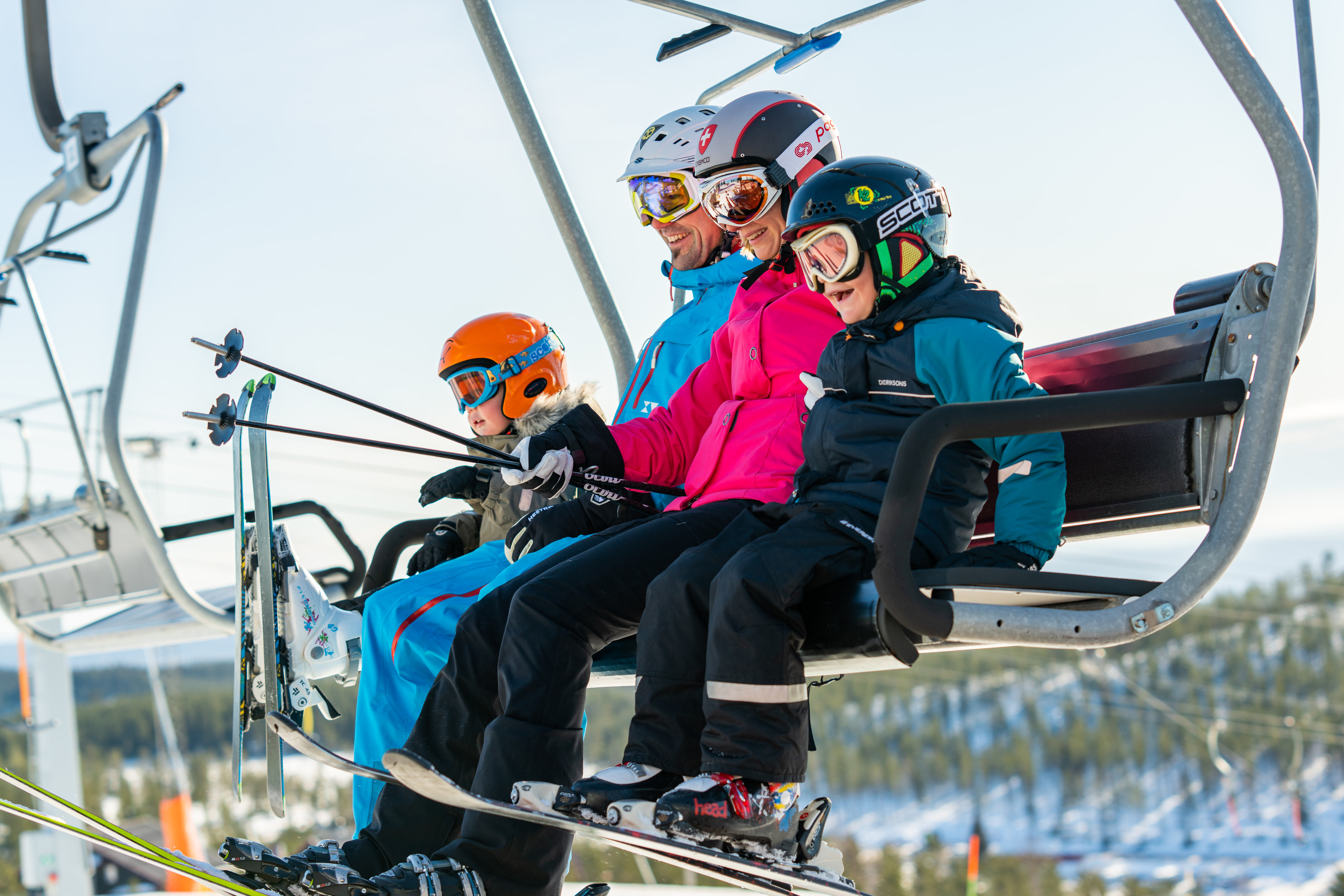 Family in colorful ski clothes riding a chairlift up the mountain on a sunny winter day.