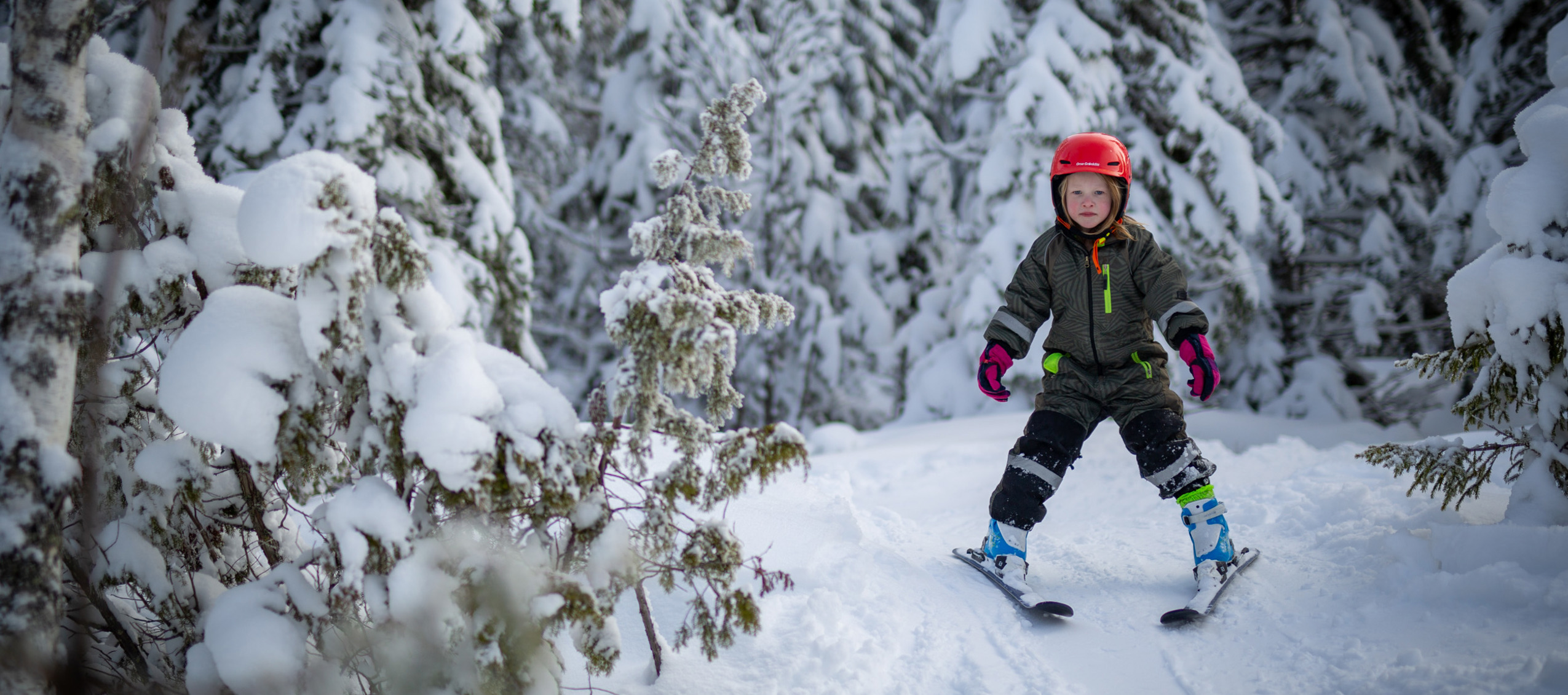 Barn med röd hjälm åker skidor på en snötäckt stig i skogen.