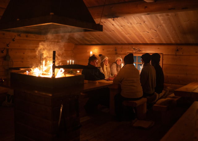 Personer sitter runt ett bord i en timrad stuga med öppen eld i mitten.