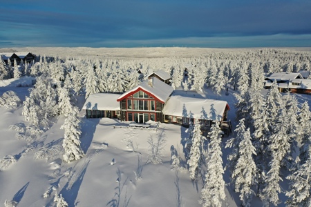 Flygbild av en byggnad omgiven av snötäckt barrskog i vinterlandskap.
