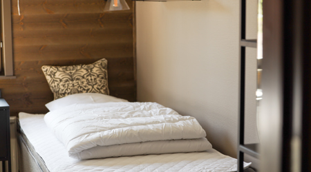 En redt enkeltseng med hvid dyne og en mønstret pude i et lyst soveværelse med træpanel.