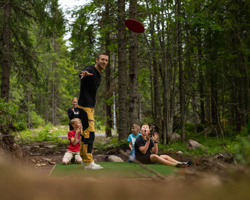 A man throws a frisbee while children and adults watch at a disc golf course in the forest.