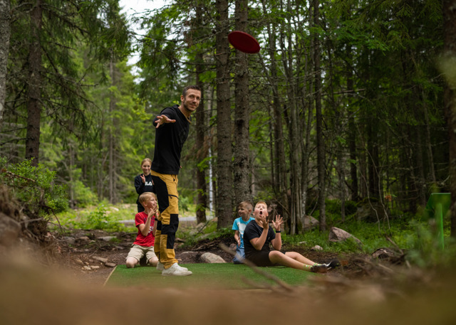 En mand kaster en frisbee, mens børn og voksne ser på i en discgolfbane i skoven.