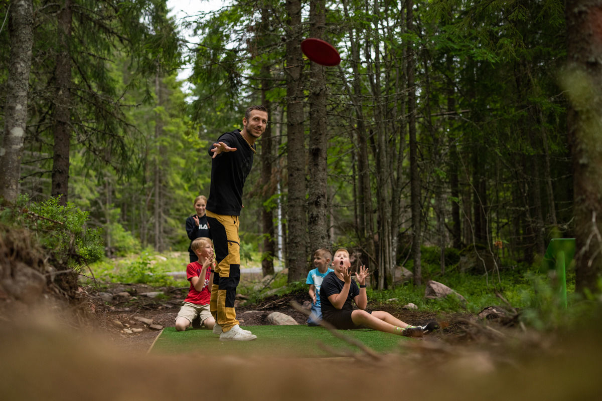 A man throws a frisbee while children and adults watch at a disc golf course in the forest.