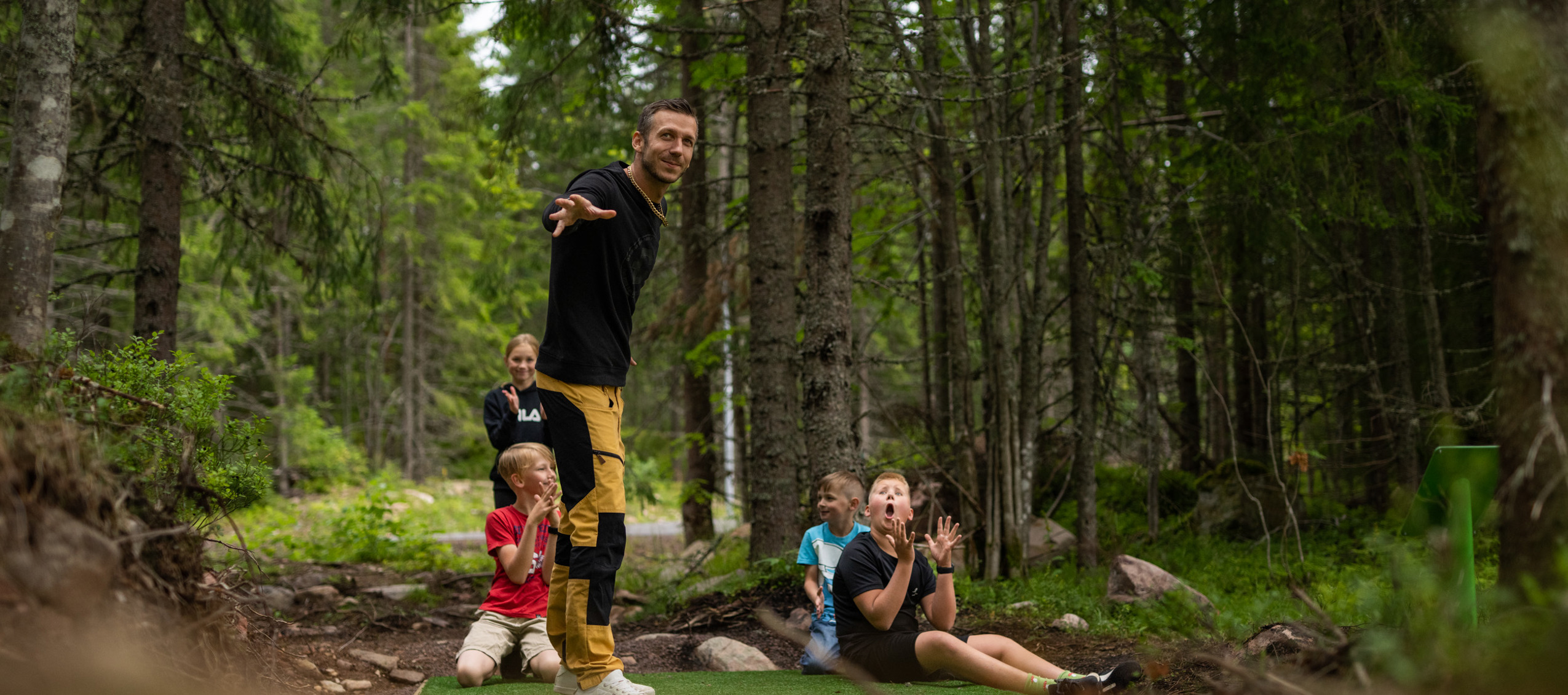 En man kastar en frisbee medan barn och vuxna ser på i en discgolfbana i skogen.
