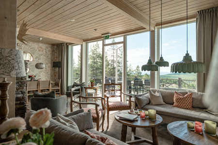 Ljus och rymlig lounge med stora panoramafönster mot skogslandskap, soffor och fåtöljer i naturtoner, samt träbord med tända ljus och dekor.
