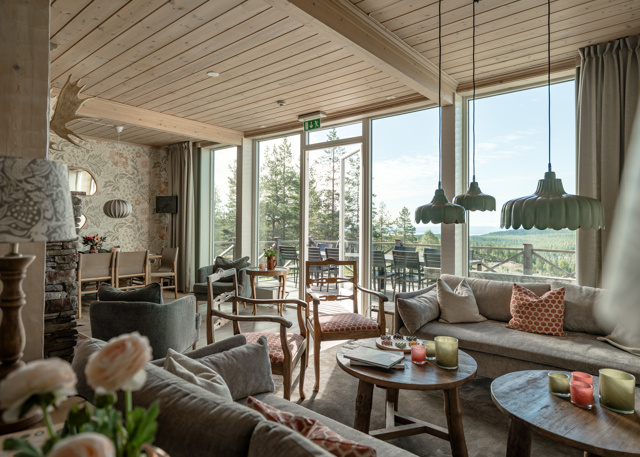 Ljus och rymlig lounge med stora panoramafönster mot skogslandskap, soffor och fåtöljer i naturtoner, samt träbord med tända ljus och dekor.
