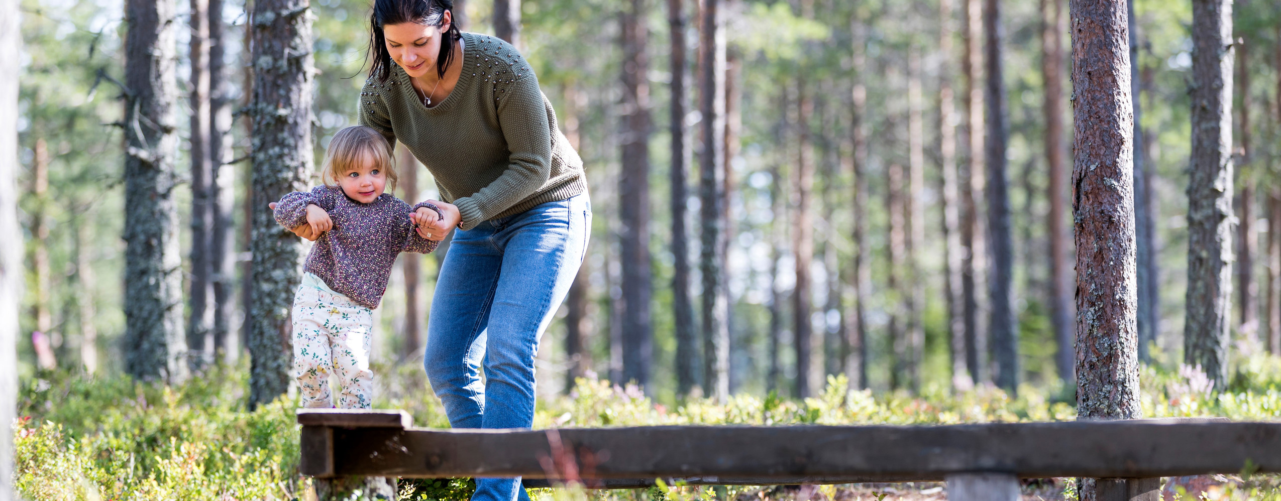 Adult and young child balancing together on a wooden beam in the forest.