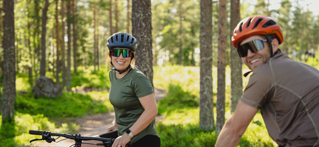 En kvinna och en man med cykelhjälmar och solglasögon pausar på en skogsstig under en cykeltur i soligt väder.