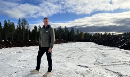 En man står på ett snötäckt område omgiven av skog under en molnig men ljus himmel. Han bär mörka byxor och en grön fleecejacka med Orsa Grönklitts logotyp.