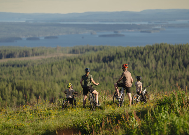Familj med cyklar pausar i en blommande sommaräng med utsikt över skogsklädda kullar och en sjö i bakgrunden.