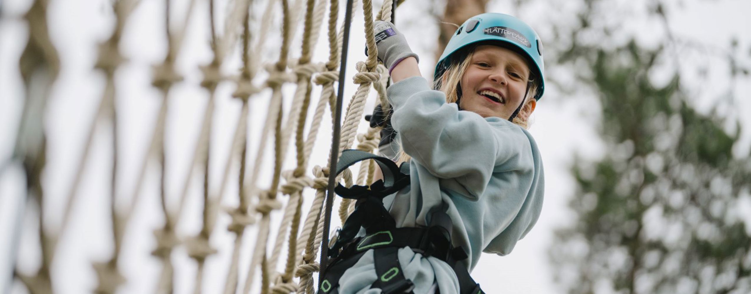 A child wearing a blue helmet and harness climbs a rope net in an adventure park. The child is smiling and looking at the camera.
