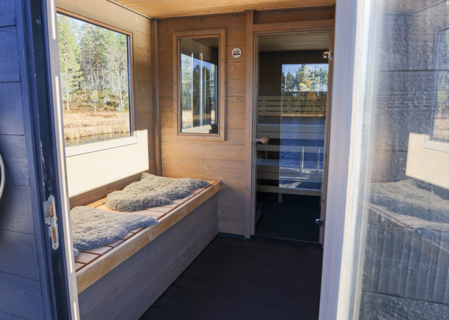 Inde fra et saunahus med vinduer mod natur og sø. Træinteriør, bænke med fåreskind og udsigt over vandet.