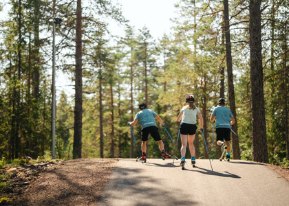 Tre ungdomar åker rullskidor uppför en asfalterad backe genom en somrig tallskog.