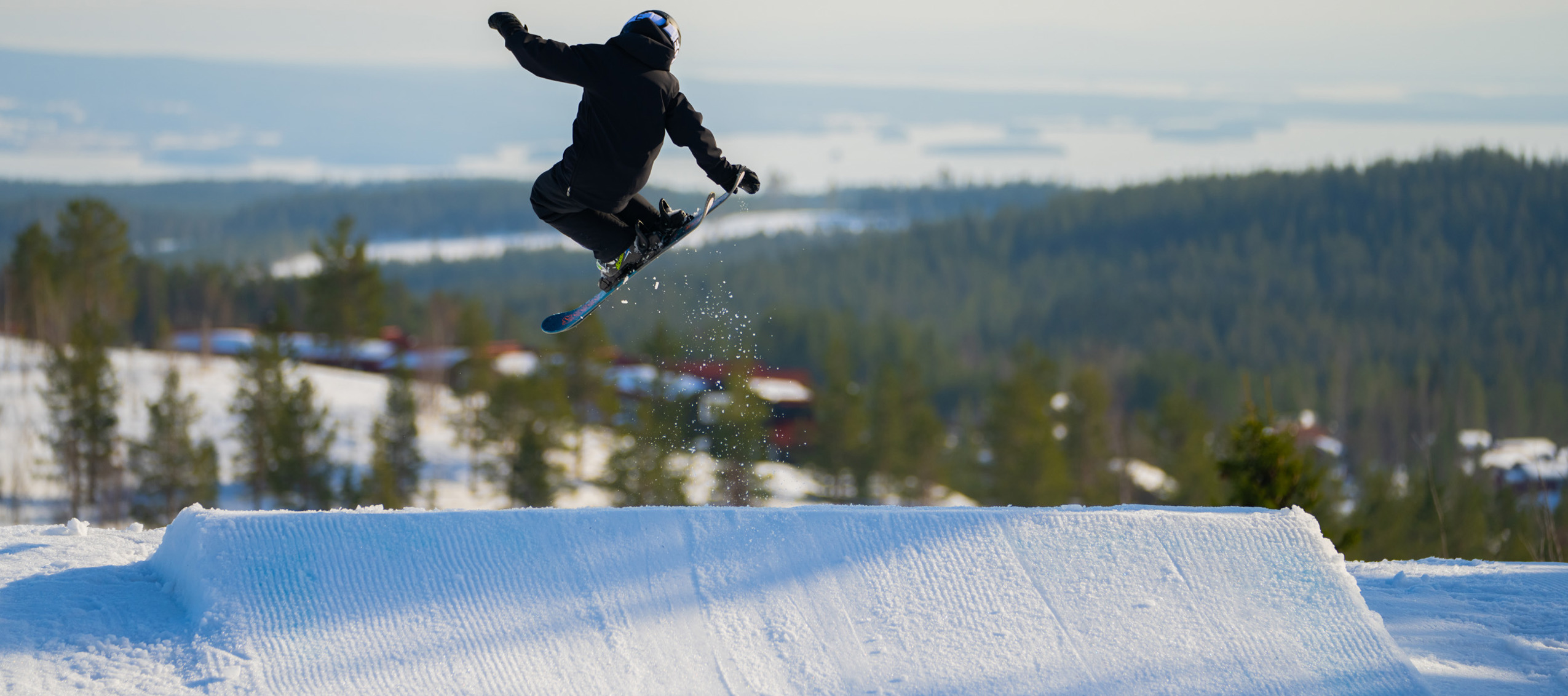 En snowboarder i sort udstyr laver et hop i snowparken med udsigt over det snedækkede landskab og hytter i baggrunden.