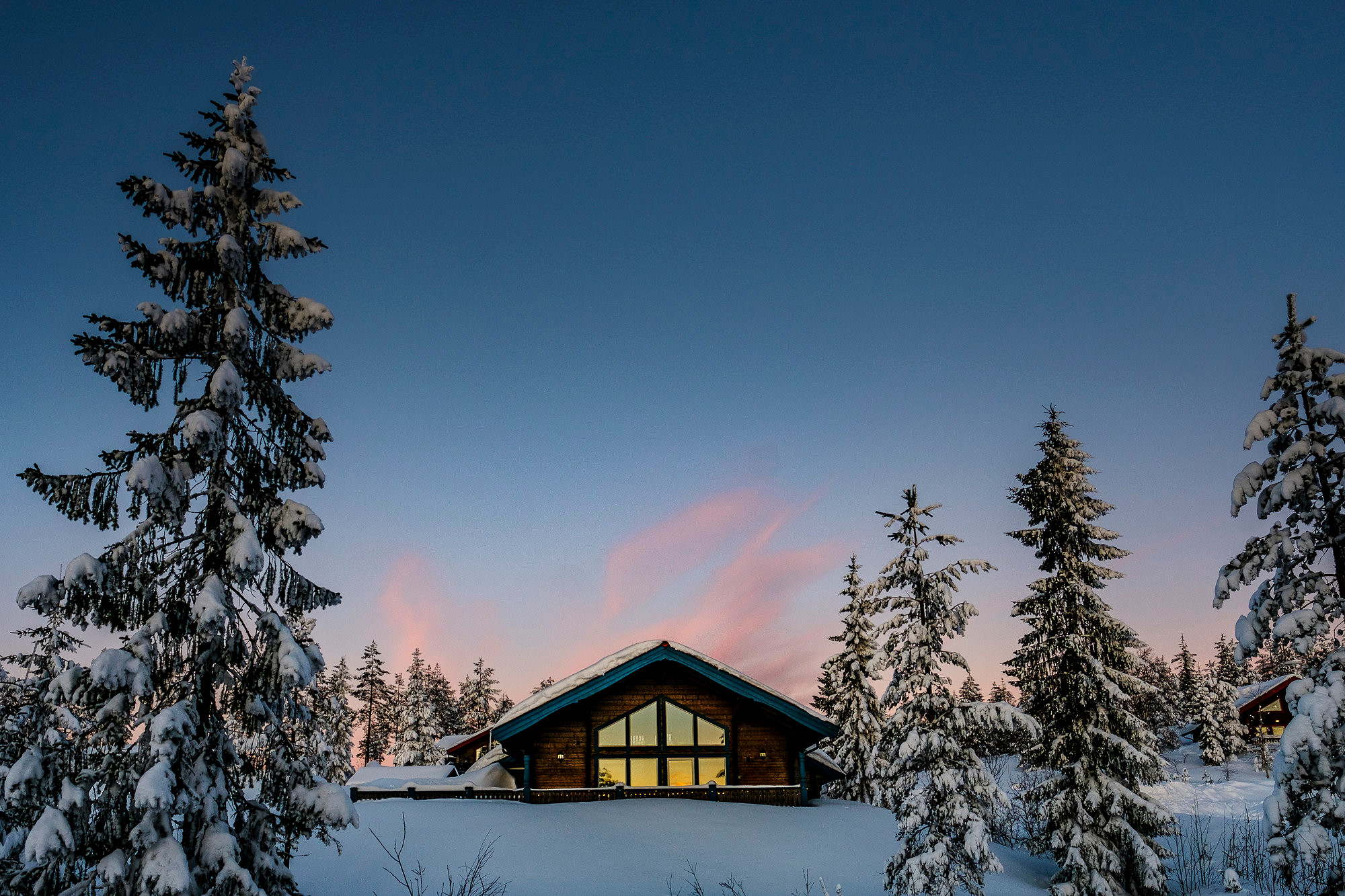 Cabin in a winter landscape at Orsa Grönklitt, surrounded by snow-covered trees with a pink sunset in the background.