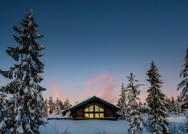 Stuga i vinterlandskap vid Orsa Grönklitt, omgiven av snötyngda träd med rosa solnedgång i bakgrunden.