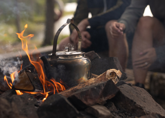Kaffepanna värms över öppen eld vid en lägerplats, med personer sittande i bakgrunden.