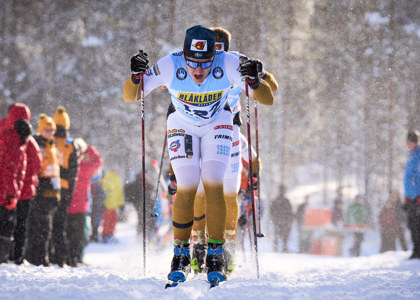 Langrendsløber i konkurrenceuniform skubber sig frem i sporet, med flere løbere bagved og publikum langs siderne i et vinterlandskab.