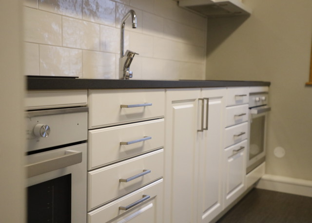 Køkken med hvide skabe, rustfri ovne og mørk bordplade.